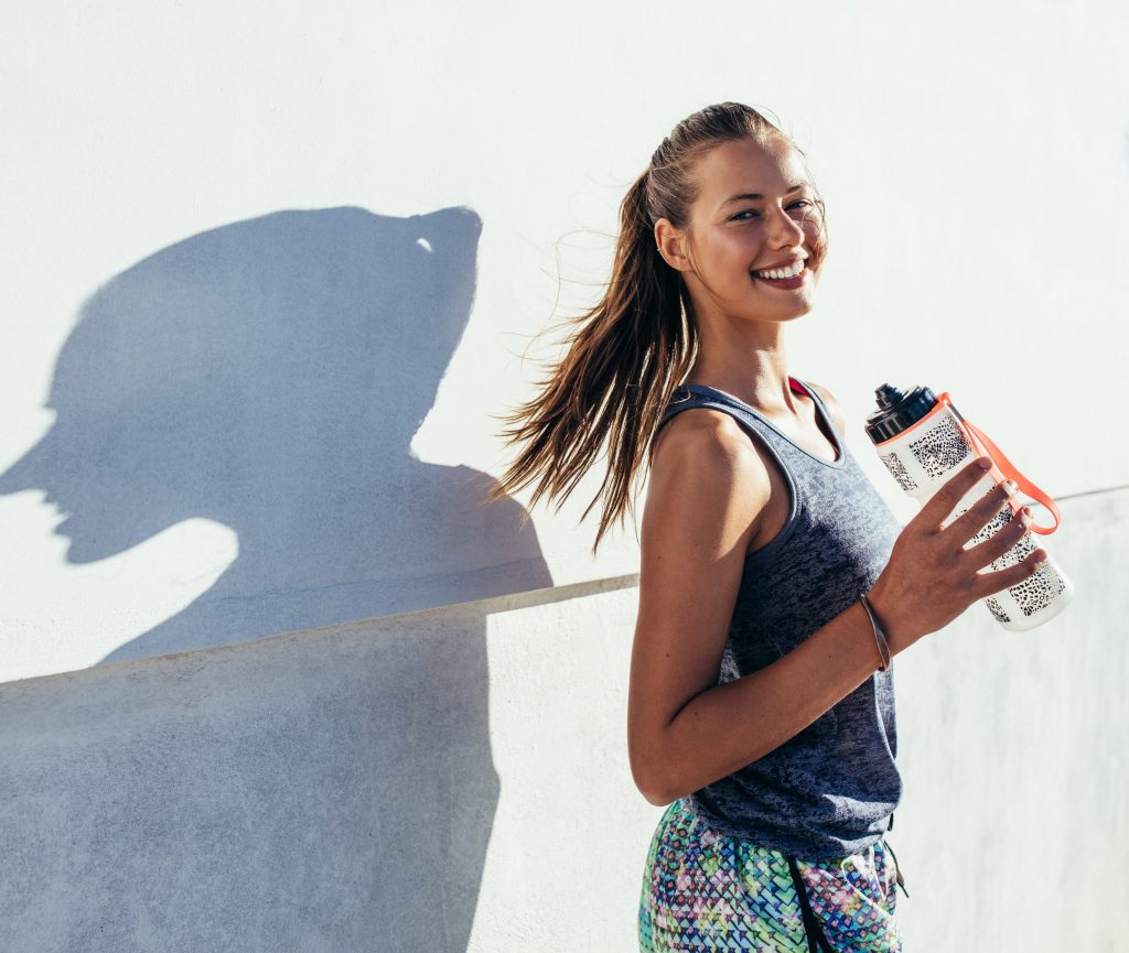 Shot of beautiful female runner standing outdoors holding water bottle. Fitness woman taking a break after running workout.